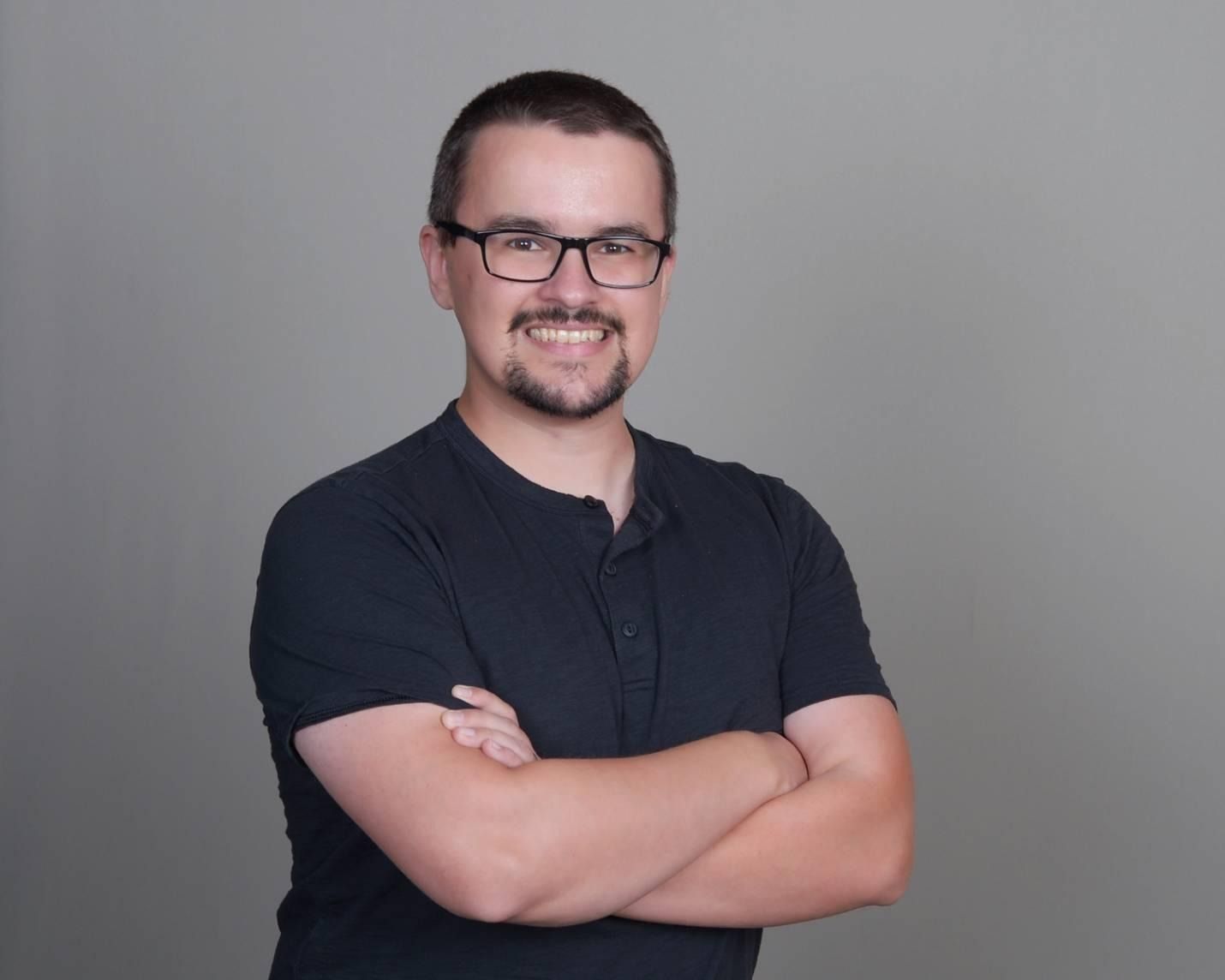 A picture of Jadon visible from the waist up. He is a man in his 30s with white, tanned skin, dark brown hair, brown eyes. He wears glasses and has a goatee beard and mustache. He is smiling with his arms crossed across his chest. Jadon wears a black shirt and stands against a gray/tan background.